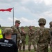 A group of military personnel in OCP (Operational Camouflage Pattern) uniforms are gathered in an outdoor field setting, appearing to receive a briefing or debriefing. A red range flag is visible flying on a pole in the background, indicating an active or recently active firing range. One civilian in a black 'Roughrider Region VI BWC' t-shirt is also present among the group.