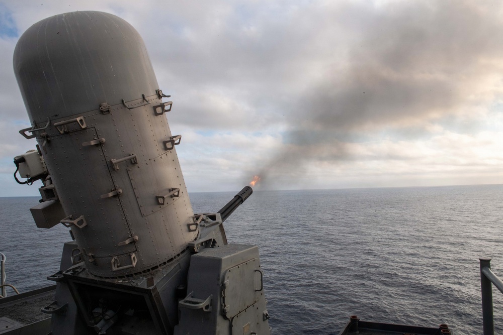 A Close-In Weapon System (CIWS), commonly known as 'Phalanx,' is actively firing from the deck of a naval warship at sea. A muzzle flash and smoke trail are visible as the multi-barrel rotary cannon discharges rounds toward the open ocean. The image was taken during what appears to be a live-fire exercise under overcast skies.