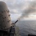 A Close-In Weapon System (CIWS), commonly known as 'Phalanx,' is actively firing from the deck of a naval warship at sea. A muzzle flash and smoke trail are visible as the multi-barrel rotary cannon discharges rounds toward the open ocean. The image was taken during what appears to be a live-fire exercise under overcast skies.