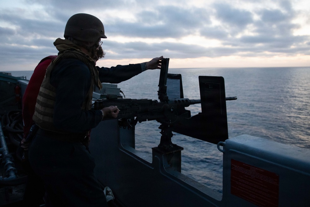 A military personnel member wearing a combat helmet, tactical vest, and glasses is operating or inspecting a mounted machine gun on the deck of a naval vessel. The scene is captured at dusk or dawn with an overcast sky and calm ocean in the background. Two display panels or shields are mounted alongside the weapon system on the ship's railing.