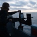 A military personnel member wearing a combat helmet, tactical vest, and glasses is operating or inspecting a mounted machine gun on the deck of a naval vessel. The scene is captured at dusk or dawn with an overcast sky and calm ocean in the background. Two display panels or shields are mounted alongside the weapon system on the ship's railing.