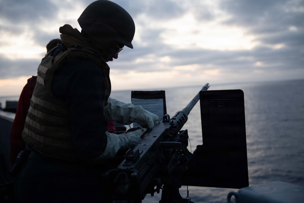 A silhouetted military personnel is working on a mounted crew-served weapon, likely a .50 caliber M2 machine gun, on the deck of a naval vessel at dusk or dawn. The figure is wearing tactical gear including a helmet, body armor vest, and white gloves. The ocean horizon and overcast sky are visible in the background.