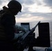 A silhouetted military personnel is working on a mounted crew-served weapon, likely a .50 caliber M2 machine gun, on the deck of a naval vessel at dusk or dawn. The figure is wearing tactical gear including a helmet, body armor vest, and white gloves. The ocean horizon and overcast sky are visible in the background.