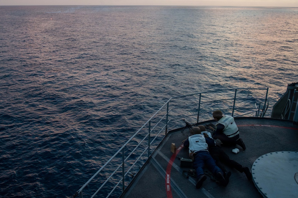 Two individuals are positioned on the bow deck of a naval or coast guard vessel at dusk, with one person prone and one kneeling, both appearing to operate or aim a mounted weapon toward the open sea. The scene is photographed from an elevated angle looking down at the forward deck. The ocean is calm with soft twilight lighting reflecting off the water.