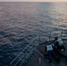 Two individuals are positioned on the bow deck of a naval or coast guard vessel at dusk, with one person prone and one kneeling, both appearing to operate or aim a mounted weapon toward the open sea. The scene is photographed from an elevated angle looking down at the forward deck. The ocean is calm with soft twilight lighting reflecting off the water.