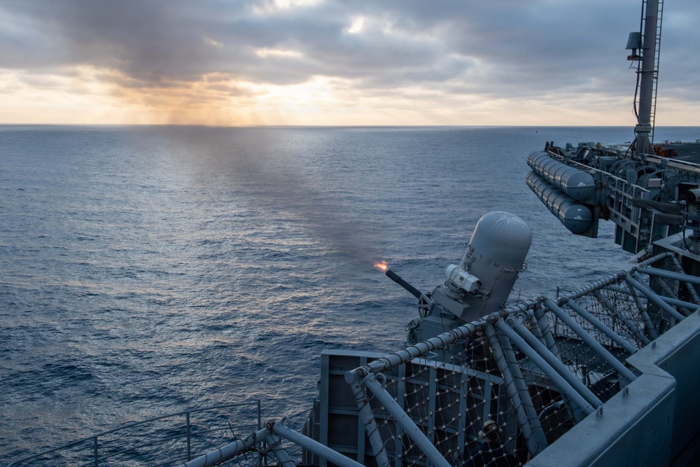 A naval vessel is conducting a live-fire exercise at sea, with a close-in weapon system (CIWS) visibly firing, indicated by the muzzle flash at the barrel tip. The scene is captured during twilight or early morning conditions with overcast skies and calm ocean waters. The ship's deck infrastructure, railings, and additional weapon systems are visible in the background.