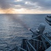A naval vessel is conducting a live-fire exercise at sea, with a close-in weapon system (CIWS) visibly firing, indicated by the muzzle flash at the barrel tip. The scene is captured during twilight or early morning conditions with overcast skies and calm ocean waters. The ship's deck infrastructure, railings, and additional weapon systems are visible in the background.
