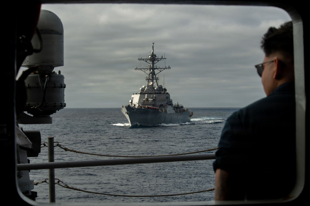 The image is taken from the deck or bridge wing of one naval vessel looking out through a rectangular opening toward a second warship at close range on open ocean. The distant vessel appears to be an Arleigh Burke-class guided-missile destroyer, identifiable by its characteristic hull shape, mast configuration, and SPY radar arrays. A sailor wearing sunglasses observes the approaching or accompanying ship from the right side of the frame.