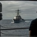 The image is taken from the deck or bridge wing of one naval vessel looking out through a rectangular opening toward a second warship at close range on open ocean. The distant vessel appears to be an Arleigh Burke-class guided-missile destroyer, identifiable by its characteristic hull shape, mast configuration, and SPY radar arrays. A sailor wearing sunglasses observes the approaching or accompanying ship from the right side of the frame.