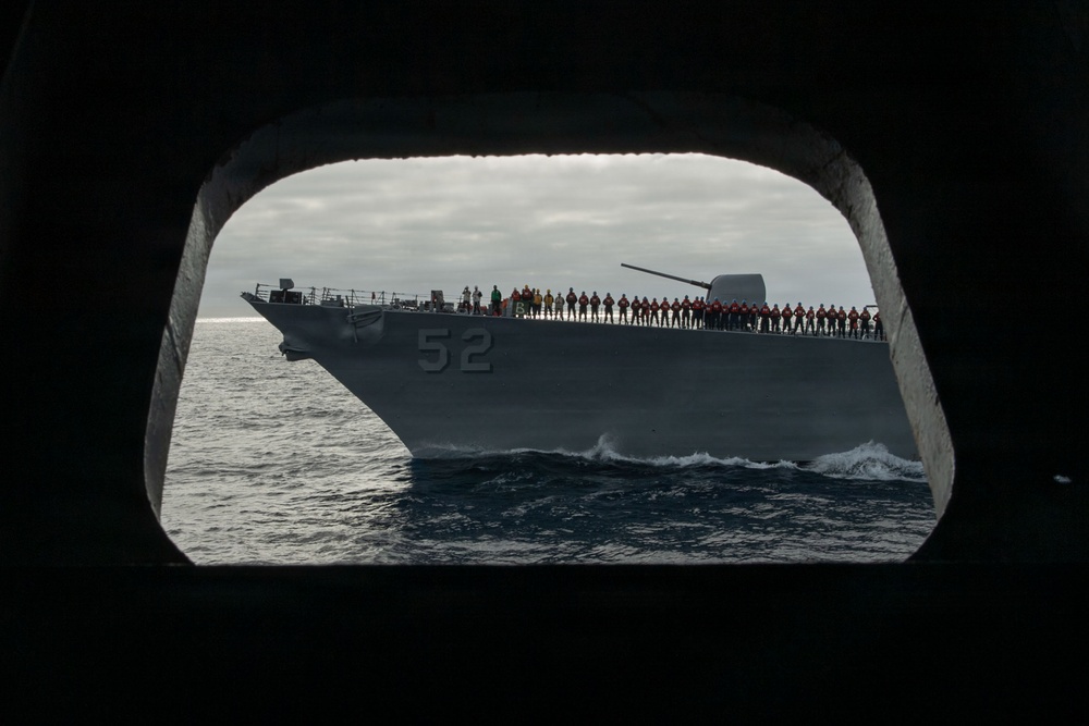 A U.S. Navy warship bearing hull number 52 is photographed through a porthole or hatch opening of another vessel at sea. The ship, likely a guided-missile cruiser or destroyer, has a large number of crew members manning the rails along its deck in a formal naval tradition. The overcast sky and choppy seas suggest open ocean conditions.