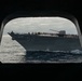 A U.S. Navy warship bearing hull number 52 is photographed through a porthole or hatch opening of another vessel at sea. The ship, likely a guided-missile cruiser or destroyer, has a large number of crew members manning the rails along its deck in a formal naval tradition. The overcast sky and choppy seas suggest open ocean conditions.