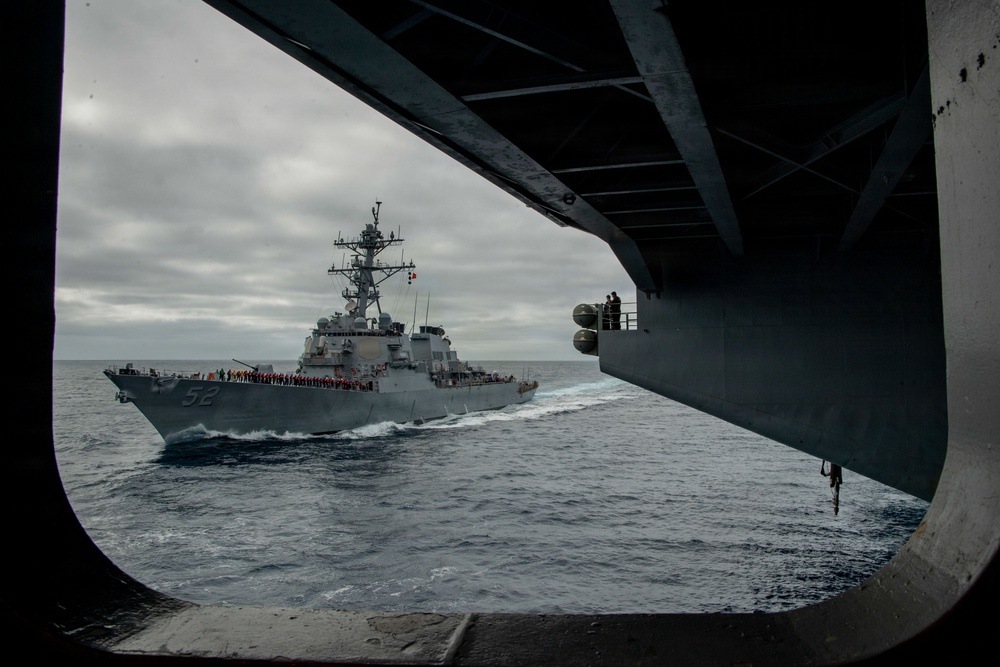 A U.S. Navy guided-missile destroyer bearing hull number 52 is photographed sailing alongside a larger vessel, viewed through a sponson or deck opening of what appears to be an aircraft carrier or large amphibious ship. The destroyer's deck is lined with crew members in colored jerseys, suggesting a formal passing or replenishment-at-sea evolution. The scene takes place in open ocean under overcast skies.