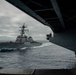 A U.S. Navy guided-missile destroyer bearing hull number 52 is photographed sailing alongside a larger vessel, viewed through a sponson or deck opening of what appears to be an aircraft carrier or large amphibious ship. The destroyer's deck is lined with crew members in colored jerseys, suggesting a formal passing or replenishment-at-sea evolution. The scene takes place in open ocean under overcast skies.
