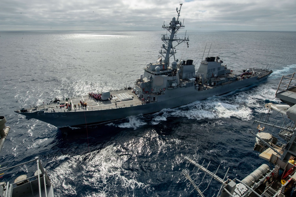 A U.S. Navy Arleigh Burke-class guided-missile destroyer is underway at sea, photographed from an elevated position on an adjacent vessel during what appears to be an underway replenishment (UNREP) or close-proximity operation. The destroyer is sailing in open ocean under overcast skies, with crew members visible on the flight deck and main deck. The image captures the ship from a slightly elevated starboard-quarter angle, showing its full profile and wake.