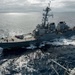 A U.S. Navy Arleigh Burke-class guided-missile destroyer is underway at sea, photographed from an elevated position on an adjacent vessel during what appears to be an underway replenishment (UNREP) or close-proximity operation. The destroyer is sailing in open ocean under overcast skies, with crew members visible on the flight deck and main deck. The image captures the ship from a slightly elevated starboard-quarter angle, showing its full profile and wake.