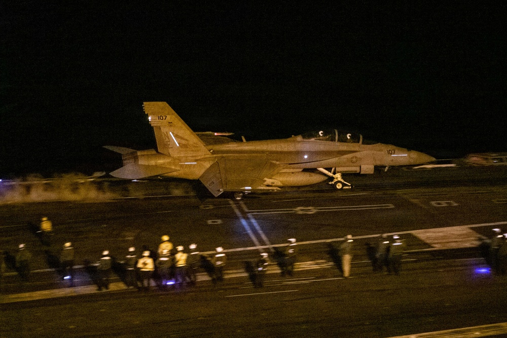 A U.S. Navy carrier-based jet aircraft, marked with the number '107', is photographed during a night flight deck operation aboard an aircraft carrier. The scene captures the aircraft either launching or taxiing on the flight deck, surrounded by flight deck crew members wearing illuminated helmets and colored jerseys. The low-light conditions and motion blur indicate this is a nighttime carrier operation.
