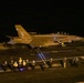 A U.S. Navy carrier-based jet aircraft, marked with the number '107', is photographed during a night flight deck operation aboard an aircraft carrier. The scene captures the aircraft either launching or taxiing on the flight deck, surrounded by flight deck crew members wearing illuminated helmets and colored jerseys. The low-light conditions and motion blur indicate this is a nighttime carrier operation.