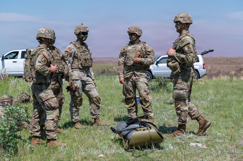 Four U.S. Army soldiers in full combat gear, including OCP (Operational Camouflage Pattern) uniforms, tactical vests, and helmets, are gathered in an open field. Their faces are painted with green camouflage face paint, suggesting a field training exercise. Civilian vehicles are parked in the background, and military duffel bags are on the ground nearby.