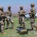 Four U.S. Army soldiers in full combat gear, including OCP (Operational Camouflage Pattern) uniforms, tactical vests, and helmets, are gathered in an open field. Their faces are painted with green camouflage face paint, suggesting a field training exercise. Civilian vehicles are parked in the background, and military duffel bags are on the ground nearby.