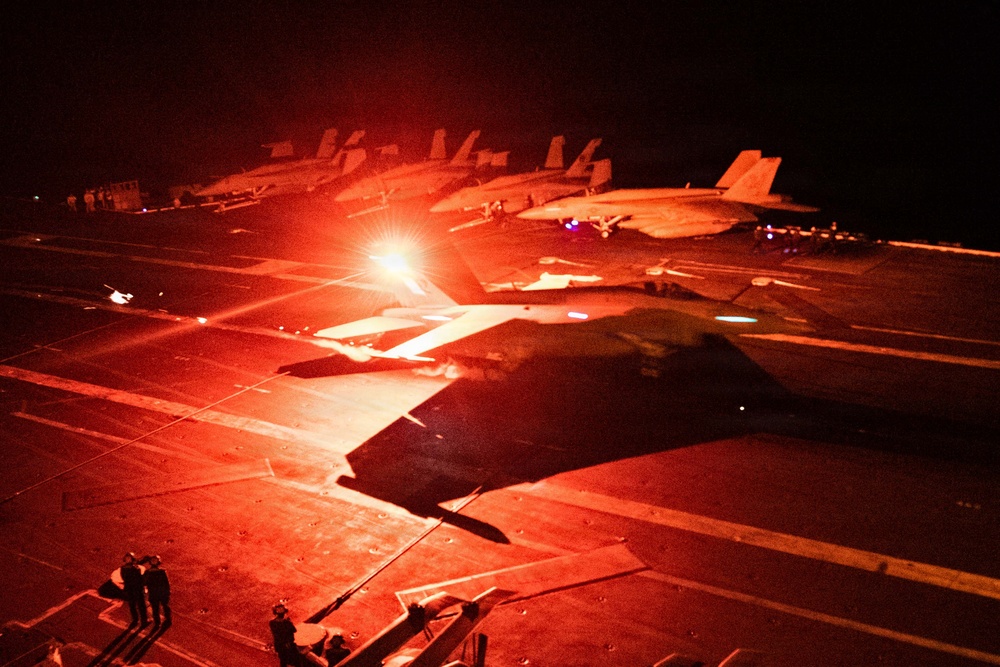 A nighttime flight deck operations scene aboard what appears to be a U.S. Navy aircraft carrier, bathed in intense red lighting. Multiple carrier-based jet aircraft, consistent with F/A-18 Hornet or Super Hornet variants, are visible on the deck with at least one appearing to be in launch position. Deck crew personnel are visible at various positions around the aircraft.