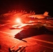 A nighttime flight deck operations scene aboard what appears to be a U.S. Navy aircraft carrier, bathed in intense red lighting. Multiple carrier-based jet aircraft, consistent with F/A-18 Hornet or Super Hornet variants, are visible on the deck with at least one appearing to be in launch position. Deck crew personnel are visible at various positions around the aircraft.