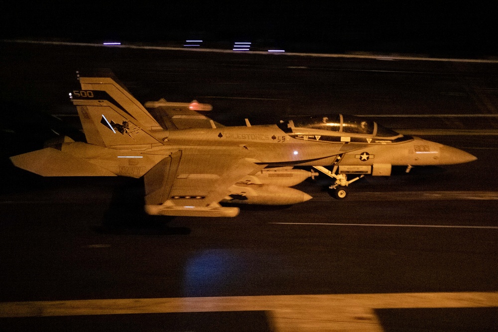 A U.S. Navy F/A-18 Super Hornet (tail number 500) is photographed at night on what appears to be an aircraft carrier flight deck. The aircraft is loaded with ordnance/external stores and appears to be taxiing or preparing for launch. The image was taken with a slow shutter speed, creating motion blur in the foreground and background.