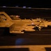 A U.S. Navy F/A-18 Super Hornet (tail number 500) is photographed at night on what appears to be an aircraft carrier flight deck. The aircraft is loaded with ordnance/external stores and appears to be taxiing or preparing for launch. The image was taken with a slow shutter speed, creating motion blur in the foreground and background.