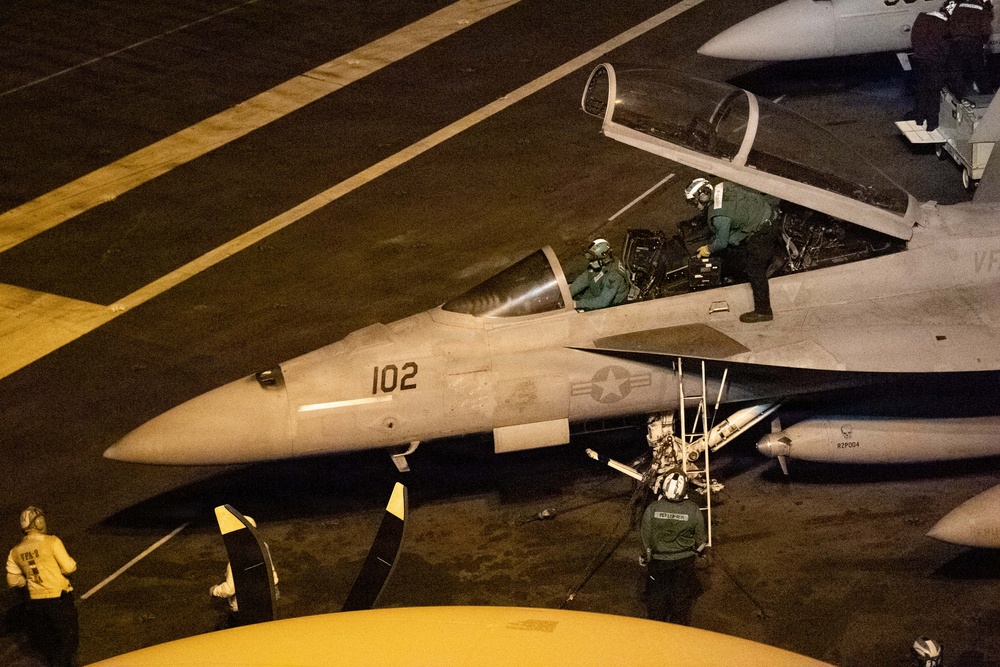 A U.S. Navy carrier-based fighter jet, marked with the number '102' and a U.S. star insignia, is being prepared on an aircraft carrier flight deck at night. At least two crew members are visible in the cockpit, with additional deck crew working around the aircraft. The scene is illuminated by artificial lighting, suggesting pre-launch or post-landing operations.