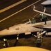 A U.S. Navy carrier-based fighter jet, marked with the number '102' and a U.S. star insignia, is being prepared on an aircraft carrier flight deck at night. At least two crew members are visible in the cockpit, with additional deck crew working around the aircraft. The scene is illuminated by artificial lighting, suggesting pre-launch or post-landing operations.
