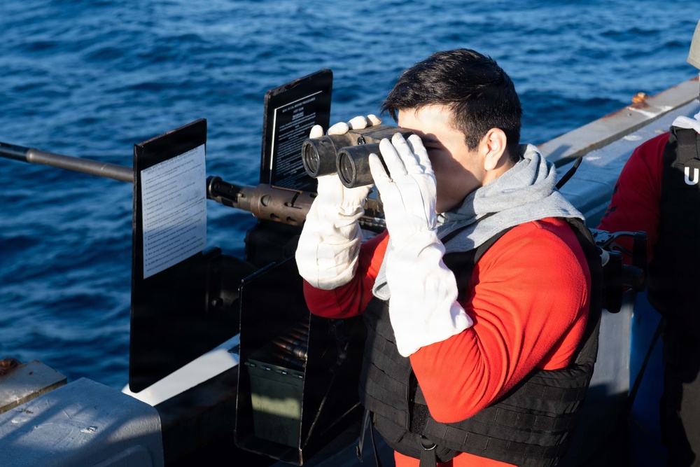 A person wearing a red long-sleeve shirt, gray hoodie, black tactical vest, and white gloves is using binoculars while standing on what appears to be a military or coast guard vessel at sea. The individual is positioned near mounted equipment, including what appears to be a weapon mount with an ammunition box and instructional placards. The ocean is visible in the background under clear conditions.