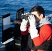 A person wearing a red long-sleeve shirt, gray hoodie, black tactical vest, and white gloves is using binoculars while standing on what appears to be a military or coast guard vessel at sea. The individual is positioned near mounted equipment, including what appears to be a weapon mount with an ammunition box and instructional placards. The ocean is visible in the background under clear conditions.