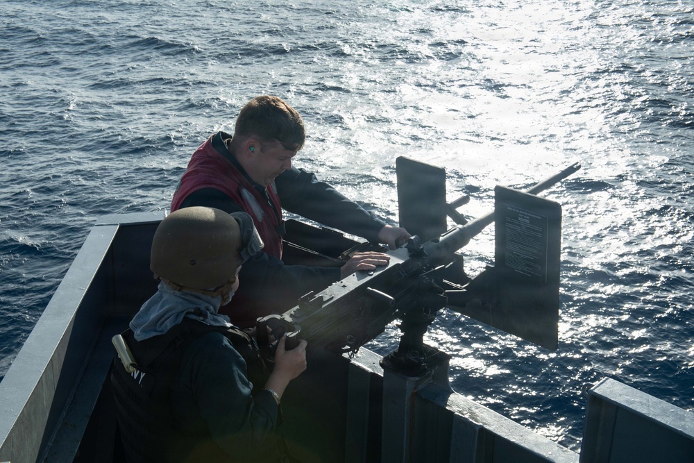 Two military personnel are working with a mounted weapon system on the deck of a naval vessel at sea. One individual is wearing a helmet and dark uniform, while the other wears a red life vest over dark clothing with ear protection. They appear to be conducting maintenance, inspection, or preparation of the weapon mount.