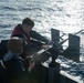Two military personnel are working with a mounted weapon system on the deck of a naval vessel at sea. One individual is wearing a helmet and dark uniform, while the other wears a red life vest over dark clothing with ear protection. They appear to be conducting maintenance, inspection, or preparation of the weapon mount.