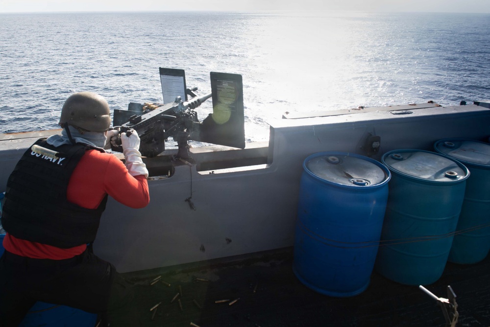 A US Navy sailor in full gear is operating a mounted machine gun on the deck of a naval vessel at sea. The sailor is wearing a helmet, face covering, gloves, a black 'US NAVY' labeled vest over a red long-sleeve shirt, and is positioned behind what appears to be an M2 .50 caliber heavy machine gun. Several blue storage drums are visible on the deck to the right, and spent brass casings can be seen on the deck surface.