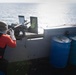 A US Navy sailor in full gear is operating a mounted machine gun on the deck of a naval vessel at sea. The sailor is wearing a helmet, face covering, gloves, a black 'US NAVY' labeled vest over a red long-sleeve shirt, and is positioned behind what appears to be an M2 .50 caliber heavy machine gun. Several blue storage drums are visible on the deck to the right, and spent brass casings can be seen on the deck surface.