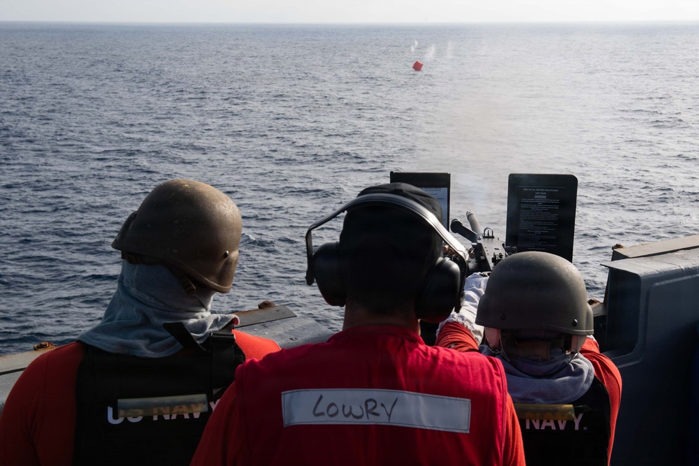 Three U.S. Navy personnel are conducting a live-fire exercise from the deck of a naval vessel at sea. The sailor in the center, identified by name tape as 'LOWRY,' is operating a mounted weapon system firing at a red target buoy visible in the water. All personnel are wearing required safety gear including helmets, hearing protection, and face coverings.