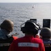 Three U.S. Navy personnel are conducting a live-fire exercise from the deck of a naval vessel at sea. The sailor in the center, identified by name tape as 'LOWRY,' is operating a mounted weapon system firing at a red target buoy visible in the water. All personnel are wearing required safety gear including helmets, hearing protection, and face coverings.