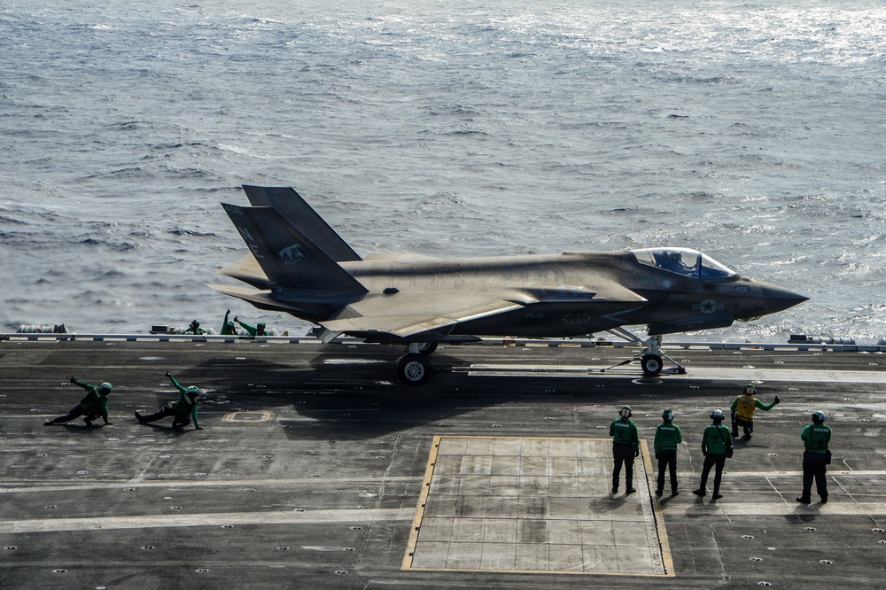An F-35C Lightning II stealth fighter jet is positioned on the flight deck of a U.S. Navy aircraft carrier, appearing to be in a pre-launch or taxi configuration. Multiple flight deck crew members in green jerseys are visible, performing launch coordination duties. The ocean is visible in the background, confirming an at-sea operational environment.