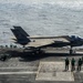 An F-35C Lightning II stealth fighter jet is positioned on the flight deck of a U.S. Navy aircraft carrier, appearing to be in a pre-launch or taxi configuration. Multiple flight deck crew members in green jerseys are visible, performing launch coordination duties. The ocean is visible in the background, confirming an at-sea operational environment.