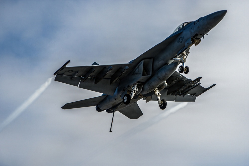 A U.S. Navy F/A-18 Super Hornet is captured in a dynamic low-altitude flight with landing gear still extended, suggesting a recent carrier catapult launch or low-speed pass. The aircraft is heavily loaded with multiple underwing stores including missiles and external fuel tanks. Vapor trails are visible streaming from the wingtips and engine exhausts, indicating high-performance flight conditions.