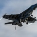 A U.S. Navy F/A-18 Super Hornet is captured in a dynamic low-altitude flight with landing gear still extended, suggesting a recent carrier catapult launch or low-speed pass. The aircraft is heavily loaded with multiple underwing stores including missiles and external fuel tanks. Vapor trails are visible streaming from the wingtips and engine exhausts, indicating high-performance flight conditions.