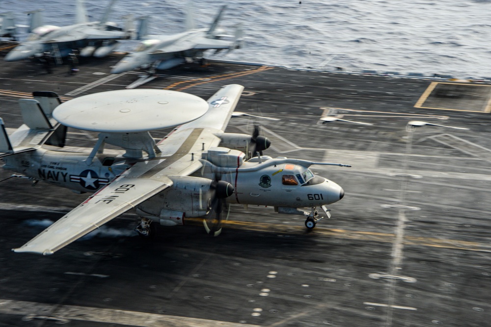 A U.S. Navy E-2 Hawkeye airborne early warning aircraft (bureau number 601) is captured mid-launch or landing on an aircraft carrier flight deck. The aircraft features its distinctive large rotodome radar dish mounted above the fuselage and twin turboprop engines. A second Navy aircraft is visible in the background on the deck.