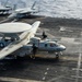 A U.S. Navy E-2 Hawkeye airborne early warning aircraft (bureau number 601) is captured mid-launch or landing on an aircraft carrier flight deck. The aircraft features its distinctive large rotodome radar dish mounted above the fuselage and twin turboprop engines. A second Navy aircraft is visible in the background on the deck.