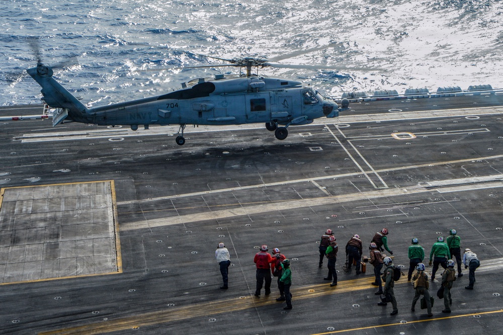 A U.S. Navy helicopter, marked '704' and labeled 'NAVY', is hovering or landing on the flight deck of an aircraft carrier at sea. Multiple deck crew members in color-coded jerseys and helmets are positioned nearby, indicating an active flight operations scenario. The ocean is visible in the background, reflecting sunlight.