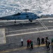 A U.S. Navy helicopter, marked '704' and labeled 'NAVY', is hovering or landing on the flight deck of an aircraft carrier at sea. Multiple deck crew members in color-coded jerseys and helmets are positioned nearby, indicating an active flight operations scenario. The ocean is visible in the background, reflecting sunlight.