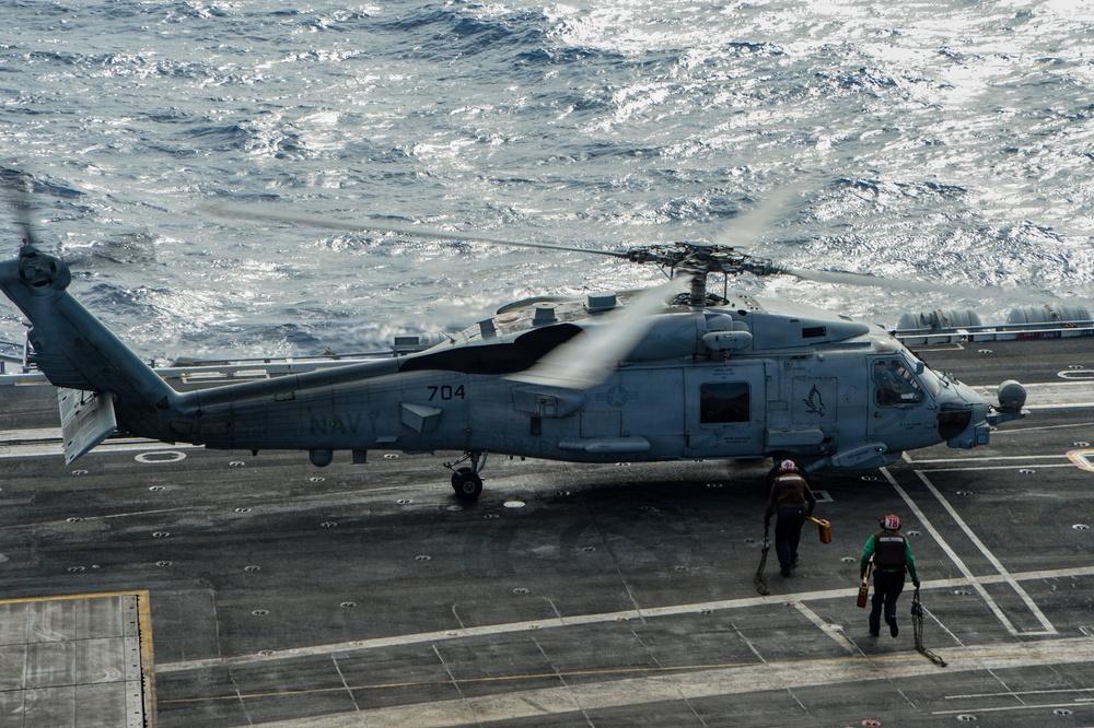 A U.S. Navy helicopter, marked '704' and labeled 'NAVY', is on the flight deck of an aircraft carrier with its rotors spinning. Two deck crew members in colored safety vests and helmets are approaching the aircraft. The ocean is visible in the background, indicating the carrier is underway at sea.