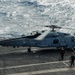 A U.S. Navy helicopter, marked '704' and labeled 'NAVY', is on the flight deck of an aircraft carrier with its rotors spinning. Two deck crew members in colored safety vests and helmets are approaching the aircraft. The ocean is visible in the background, indicating the carrier is underway at sea.