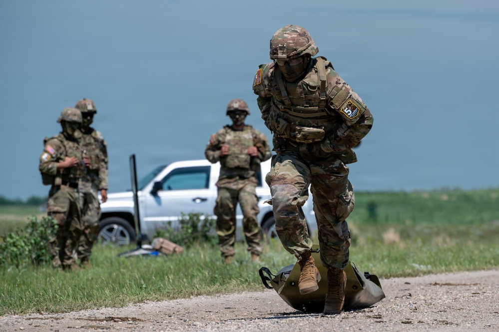 Three soldiers in U.S. Army Operational Camouflage Pattern (OCP) uniforms are engaged in what appears to be a field training exercise on a gravel road surrounded by open grassland. The foreground soldier is dragging or manipulating a heavy green duffel/kit bag on the ground, while two other soldiers stand in the background near a white pickup truck. All personnel are wearing full combat gear including helmets, body armor, and tactical vests.
