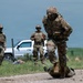 Three soldiers in U.S. Army Operational Camouflage Pattern (OCP) uniforms are engaged in what appears to be a field training exercise on a gravel road surrounded by open grassland. The foreground soldier is dragging or manipulating a heavy green duffel/kit bag on the ground, while two other soldiers stand in the background near a white pickup truck. All personnel are wearing full combat gear including helmets, body armor, and tactical vests.