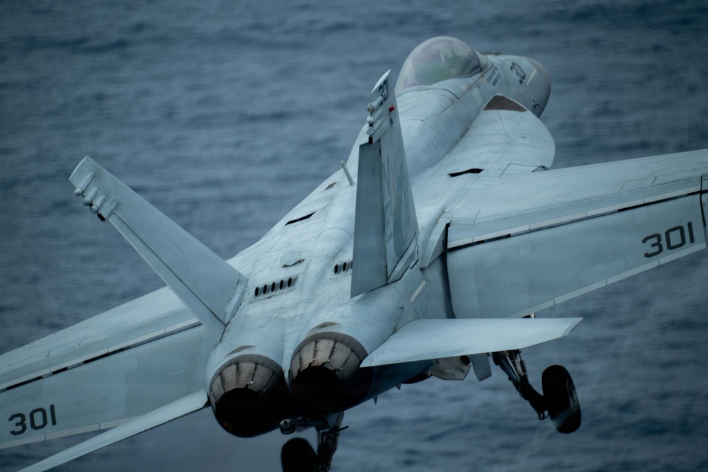 A U.S. Navy carrier-based jet aircraft, marked with the modex number '301', is captured in close-up from a rear-quarter angle, appearing to be in the process of launching from or recovering aboard an aircraft carrier over open ocean. The aircraft displays twin engines, twin tail fins, and deployed landing gear, consistent with a Boeing F/A-18E/F Super Hornet configuration. The ocean is visible in the background, indicating the vessel is at sea.